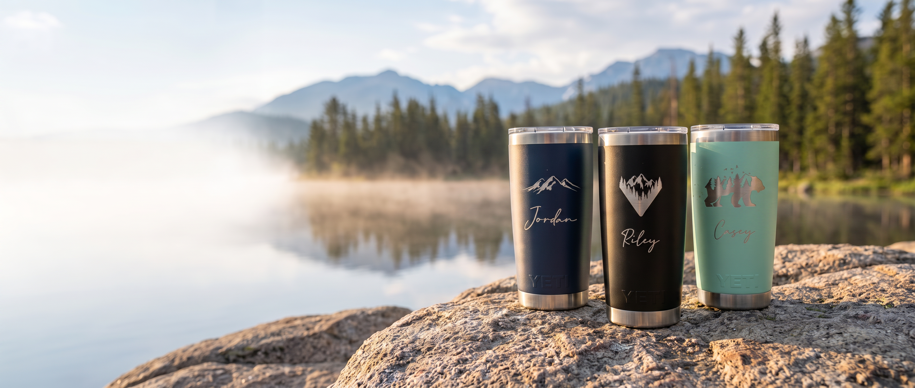 Three personalized laser-engraved YETI Rambler tumblers on a rock beside a misty alpine lake at golden hour